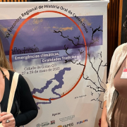 Fotografia de duas mulheres em pé, lado a lado, com os rostos desfocados, posicionadas em frente a um banner vertical. O banner anuncia o XVI Encontro Regional de História Oral do Centro-Oeste, com o tema “Emergências climáticas, Oralidades Tradicionais”, realizado na Cidade de Goiás (GO), entre os dias 27 e 29 de maio de 2025. A arte do evento é assinada por Carla Ayê. Na parte inferior do banner, constam os logotipos das instituições apoiadoras: Goiás 200 anos, FAPEG e FUNAPE.